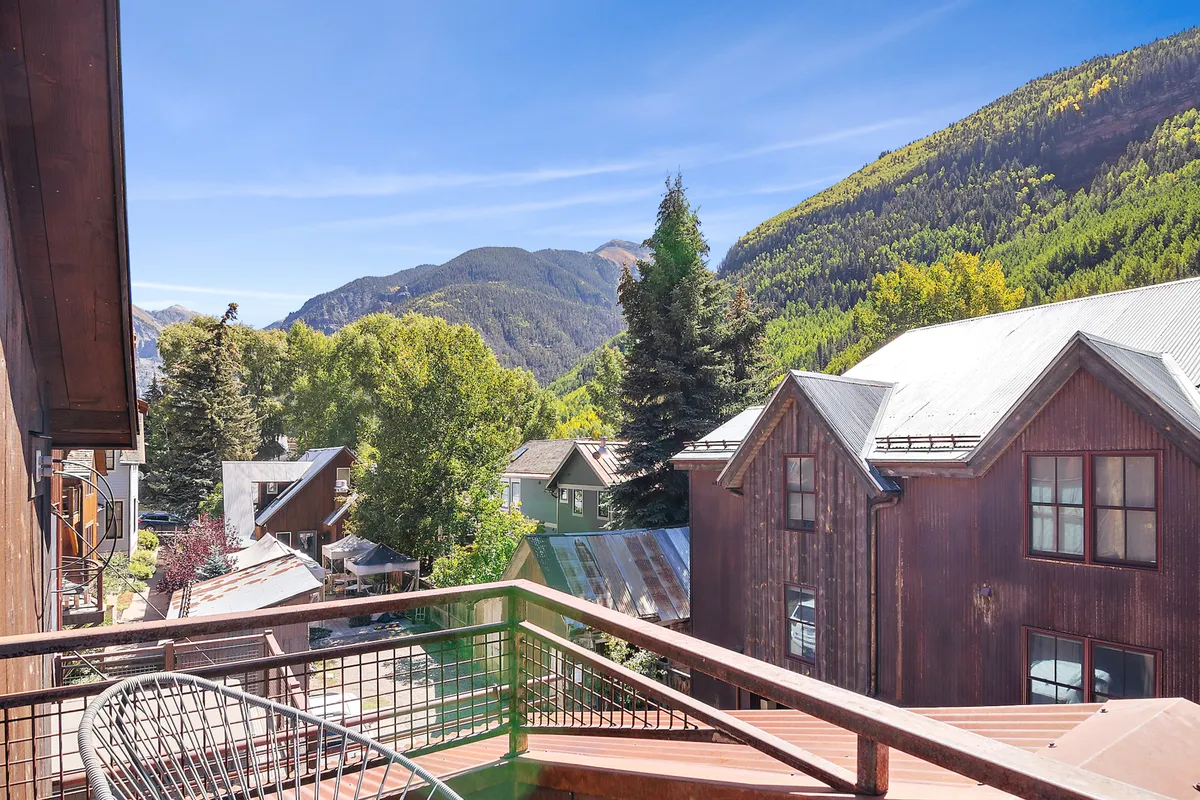 A balcony framed by a railing overlooks a dramatic mountain landscape, vibrant autumn trees, and rustic buildings. The morning sunlight enhances the peaceful ambiance, ideal for enjoying coffee or simply soaking in the views.