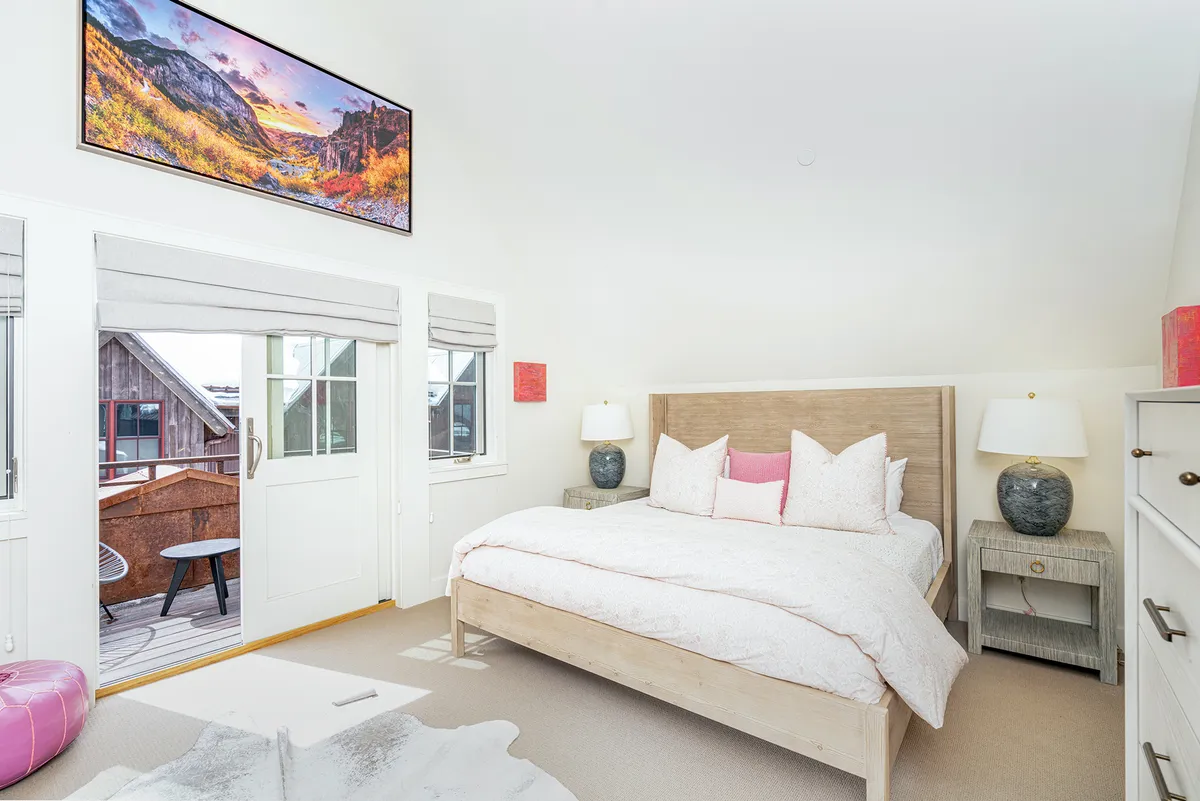 A spacious primary bedroom with a large bed adorned with decorative pillows, an art piece above the headboard, and balcony doors that open to the outside. The room is softly lit and furnished for comfort, showcasing the inviting atmosphere of a restful retreat.