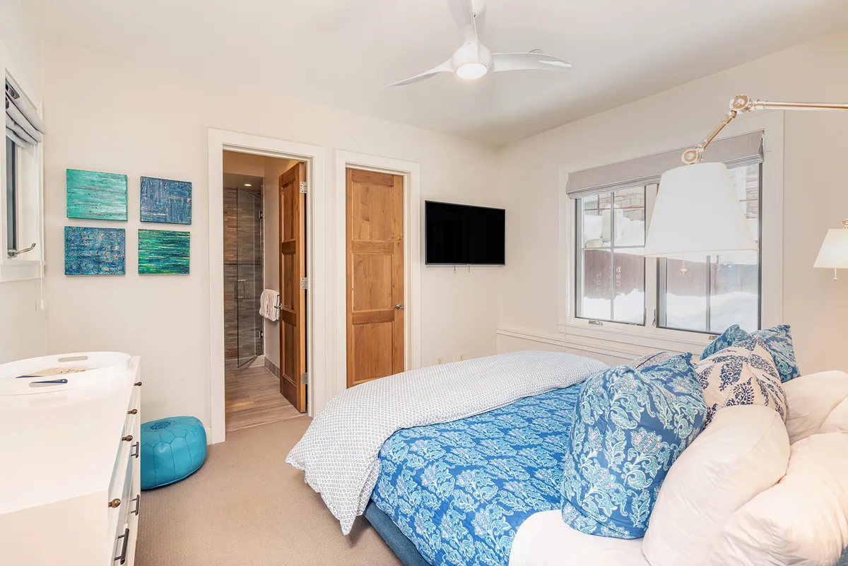 A tranquil bedroom featuring a bed with soft blue-patterned bedding, green and blue wall art, a ceiling fan, and a television. The space exudes a serene, cozy ambiance, ideal for relaxation and restful family stays.