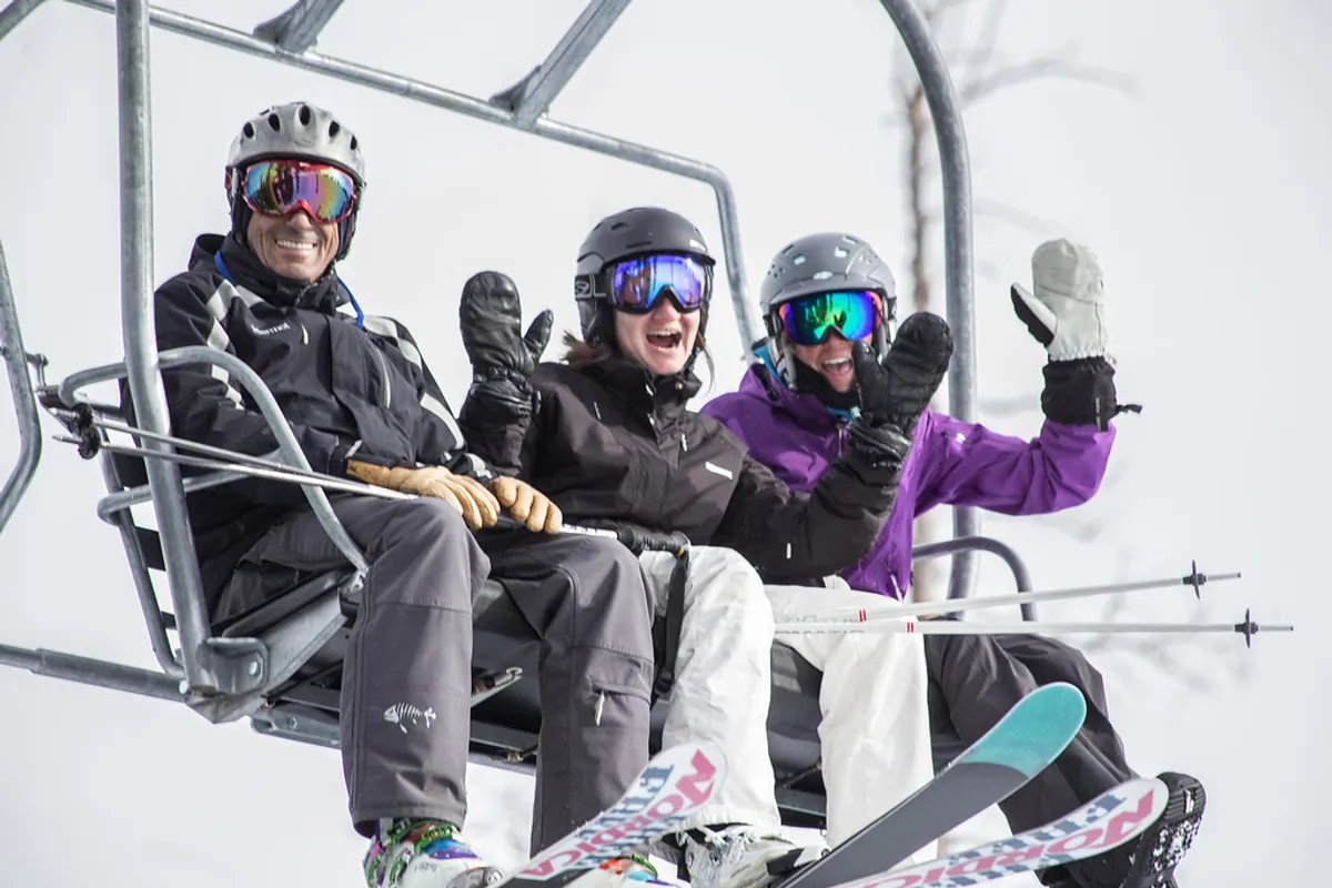 Three skiers riding a chairlift over a snowy landscape, with ski equipment visible and panoramic mountain views in the background. The mood is joyful and energetic, capturing the excitement of a day on the slopes with friends.
