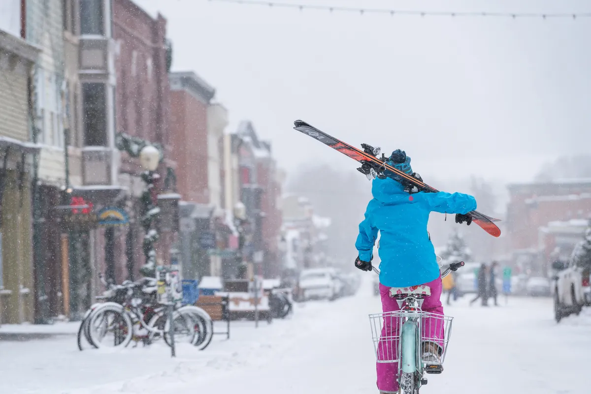 A snow-covered street lined with colorful storefronts, with a person cycling with skis, captures the playful spirit of downtown Telluride. The lively ambiance hints at vibrant local shops, winter activities, and a friendly, energetic community.