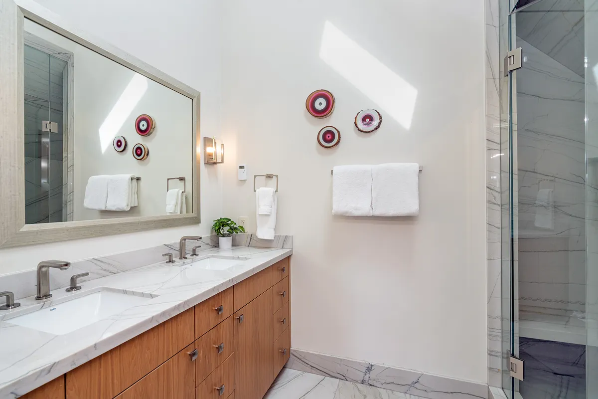 A stylish bathroom featuring double sinks on a marble countertop, modern light fixtures, decorative wall art, and neatly arranged towels. The space feels refreshing and luxurious, ideal for self-care routines and relaxation.