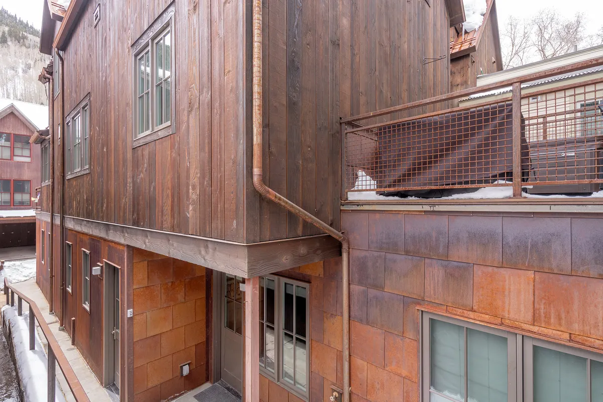 A cozy exterior view of a wooden-sided townhome with a rust-colored balcony and grill, surrounded by a snowy landscape. The inviting atmosphere speaks to outdoor relaxation and togetherness after a day of exploring.