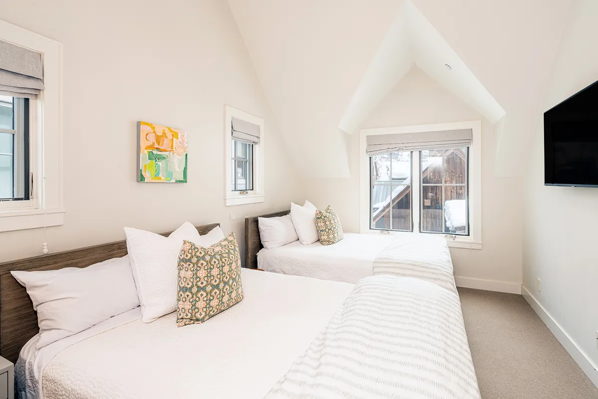 A third bedroom setting with two neatly made beds, colorful artwork on the wall, large windows that fill the space with natural light, and a television mounted on the wall. The room appears bright, comfortable, and ideal for families or friends.