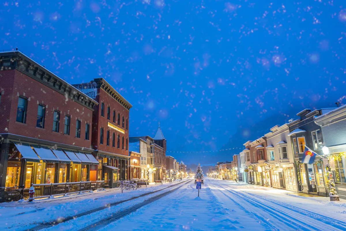 A picturesque winter scene of Telluride’s historic brick buildings on a snow-covered street, with a decorated Christmas tree and softly lit storefronts inviting families to stroll and explore. The atmosphere is festive and welcoming, perfect for a family outing.