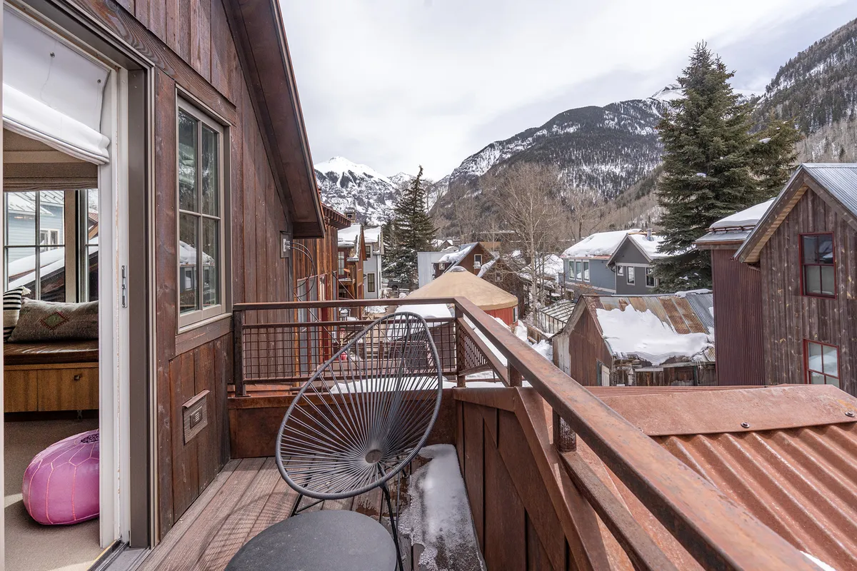 A serene balcony setup features comfortable seating facing out towards a panoramic mountain view. The surrounding architecture blends modern and rustic elements, and the tranquil outdoor atmosphere invites relaxation and socializing.