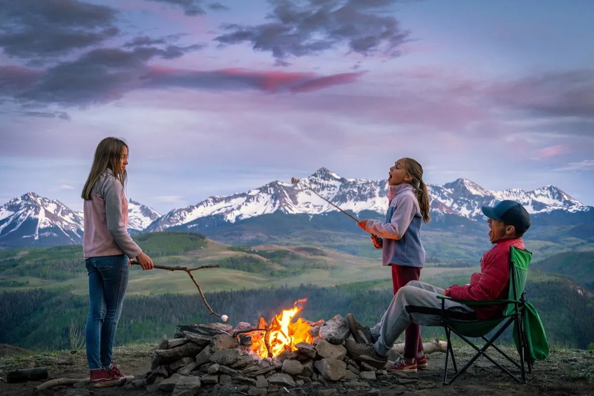 A joyful family gathers around a glowing campfire, with children roasting marshmallows and a mountain backdrop behind them. Laughter and the warmth of the fire create a sense of togetherness and outdoor adventure, set against Telluride's natural beauty.