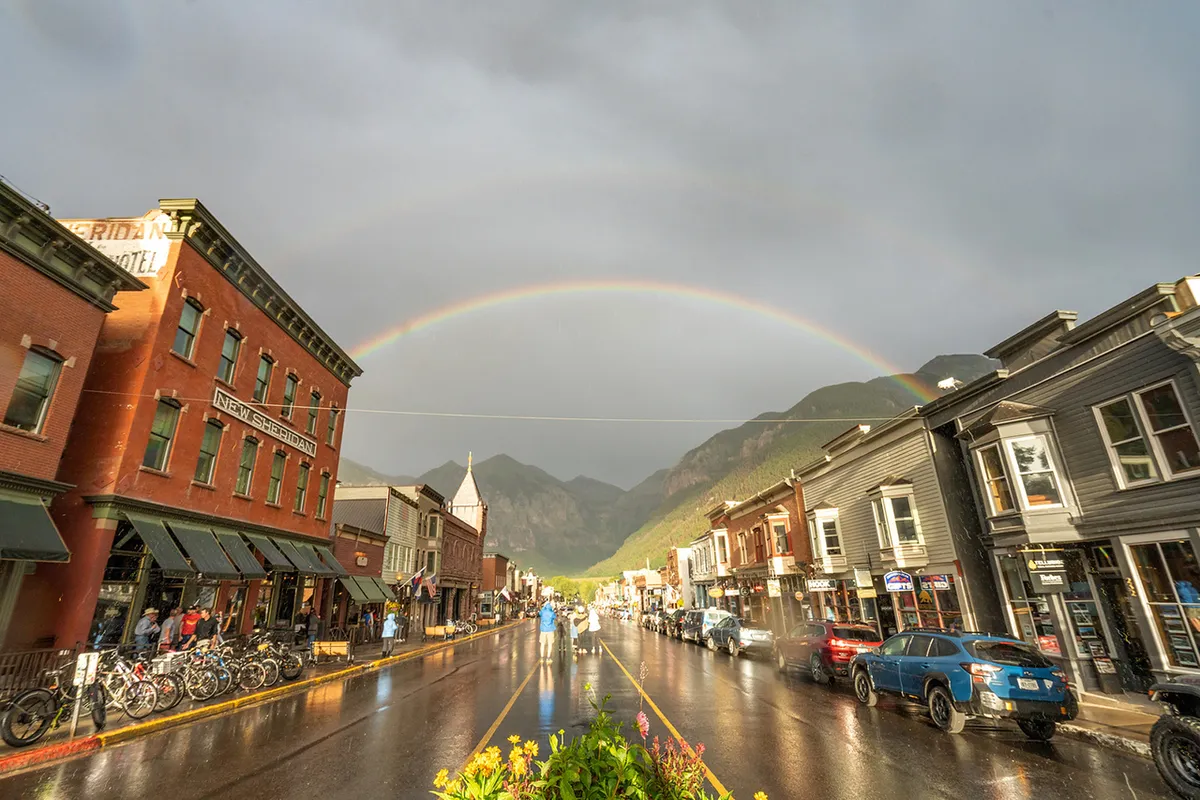A lively street scene featuring Telluride’s historic buildings, a rainbow arching overhead, wet street reflections, and people strolling and enjoying the outdoors. Bicycles and outdoor dining showcase the vibrant, walkable atmosphere of Main Street.