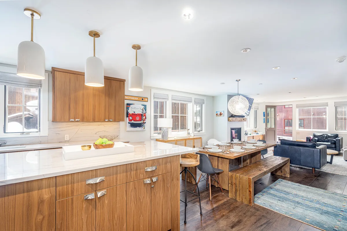 This image provides a wide view of the open-concept area, featuring the kitchen island, a dining table for eight, large windows that flood the space with sunlight, and a comfortable sofa. The scene captures the seamless flow between cooking, dining, and relaxing spaces, accented by modern furnishings and inviting natural light.